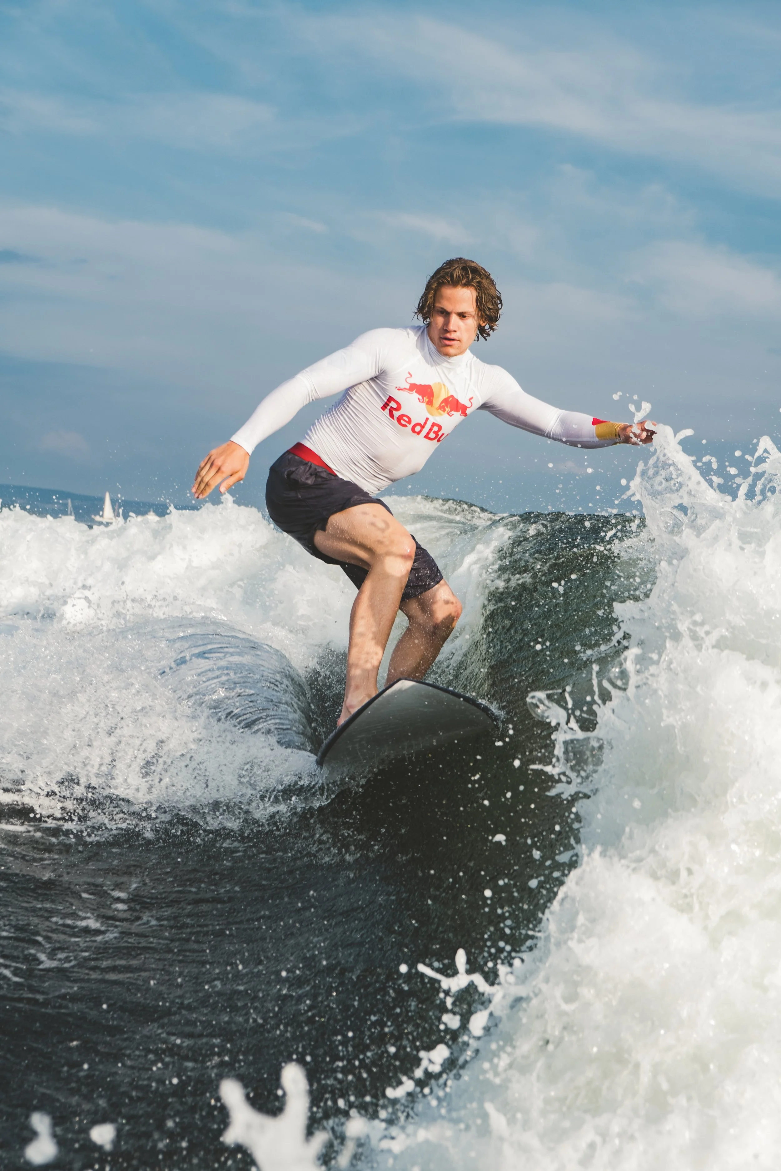 A man wearing a white Red Bull shirt and black shorts surfing on a wave.