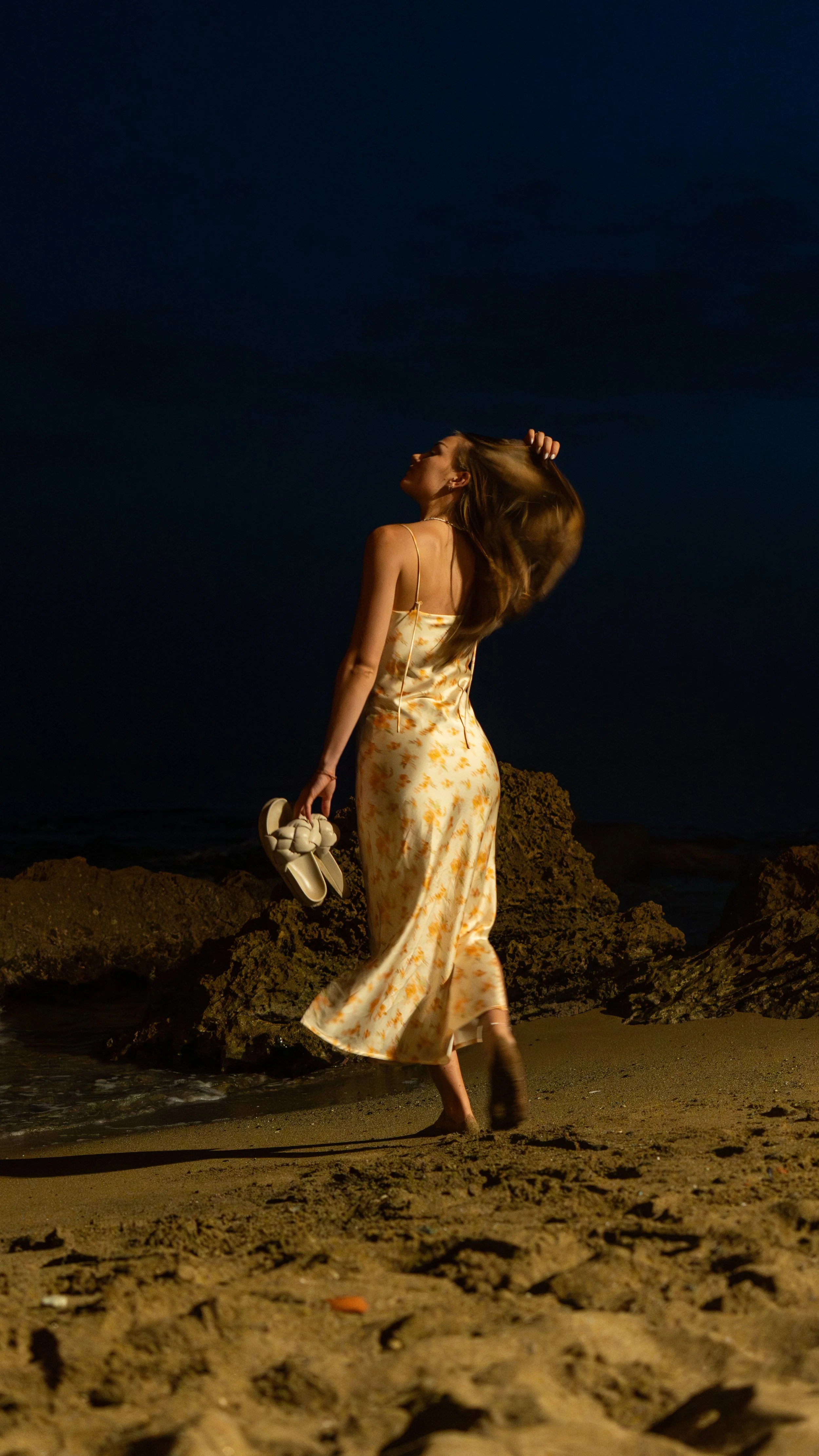 A woman in a floral dress walking barefoot on the beach at night, holding shoes, with rocks and sand in the foreground and dark sky above.
