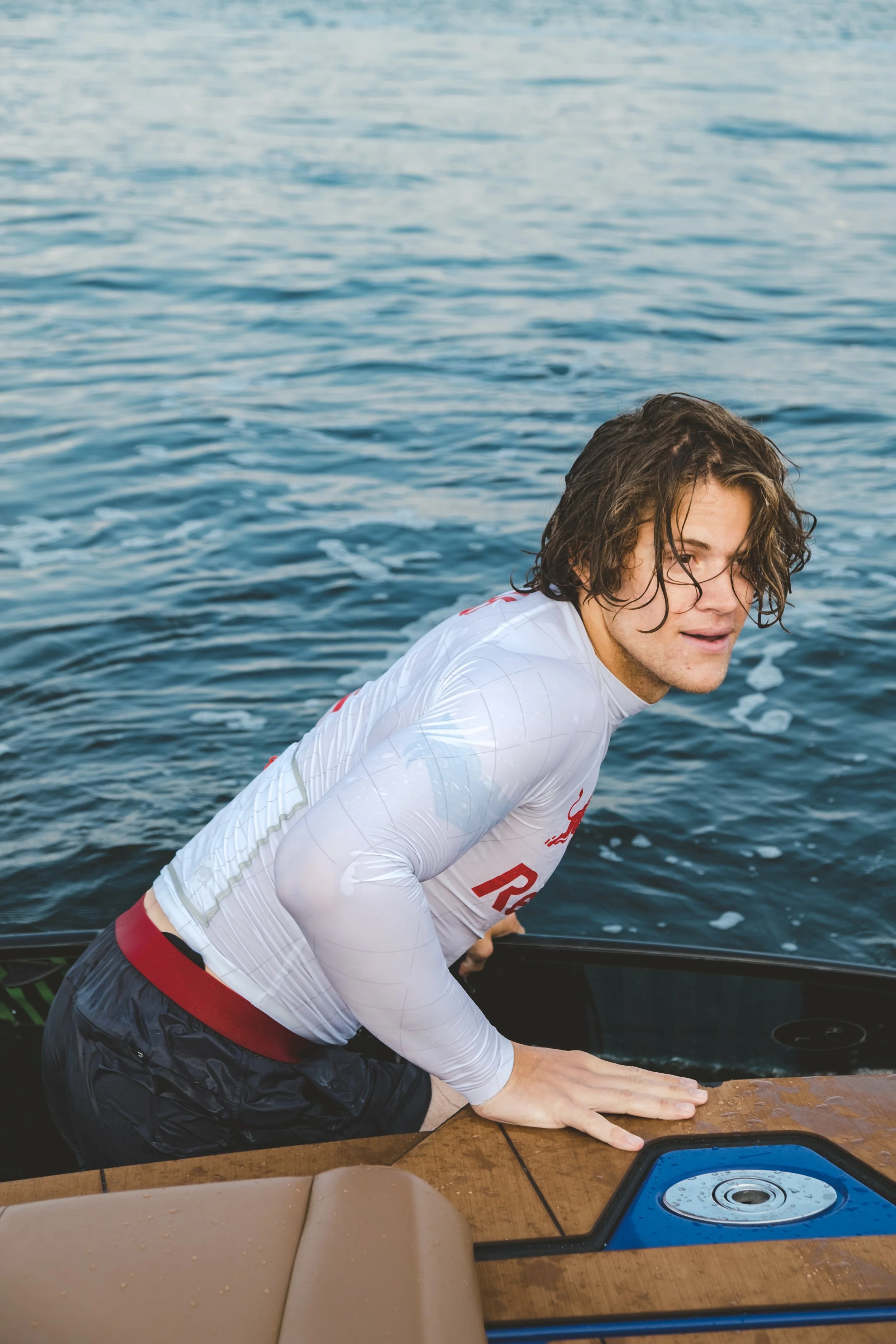A young man with wet hair wearing a white sports jersey and black shorts is leaning on a boat's wooden surface near the water.