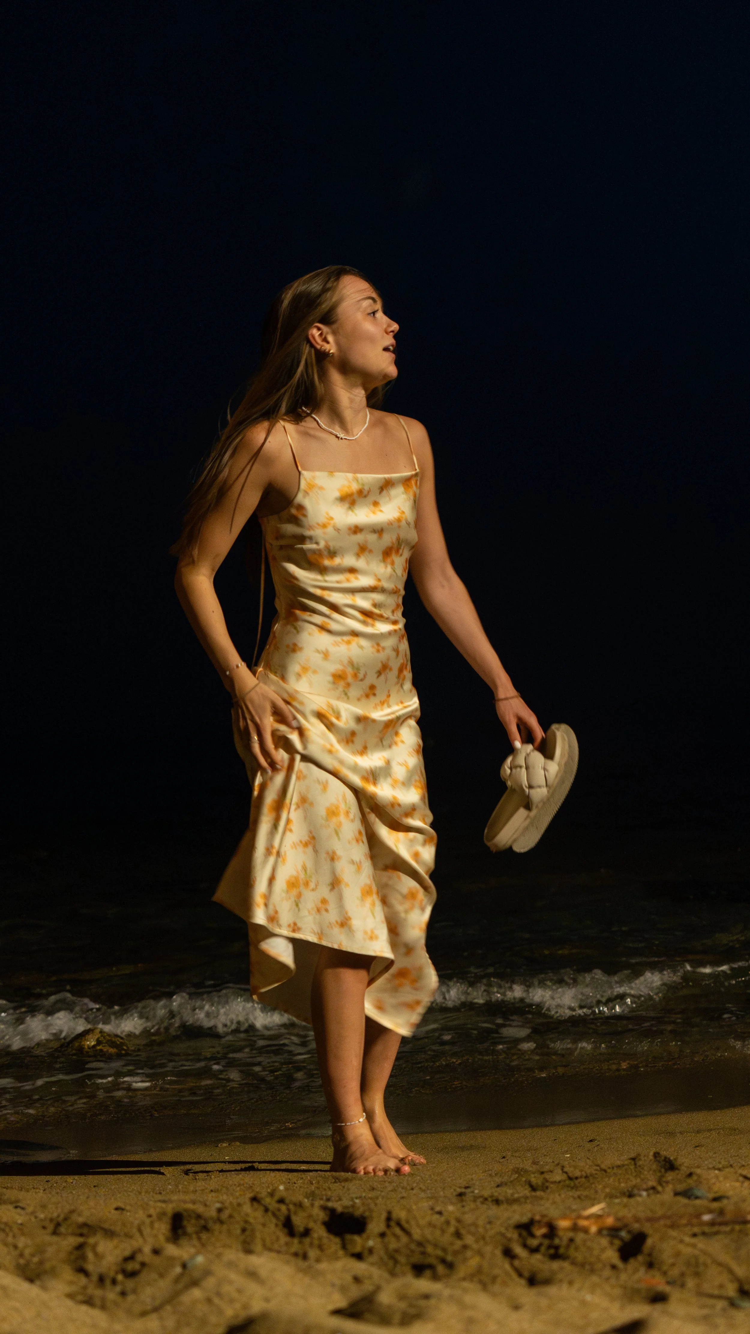 A woman walking barefoot on the beach at night, holding her shoes in one hand, wearing a floral dress, with the dark ocean and sky in the background.