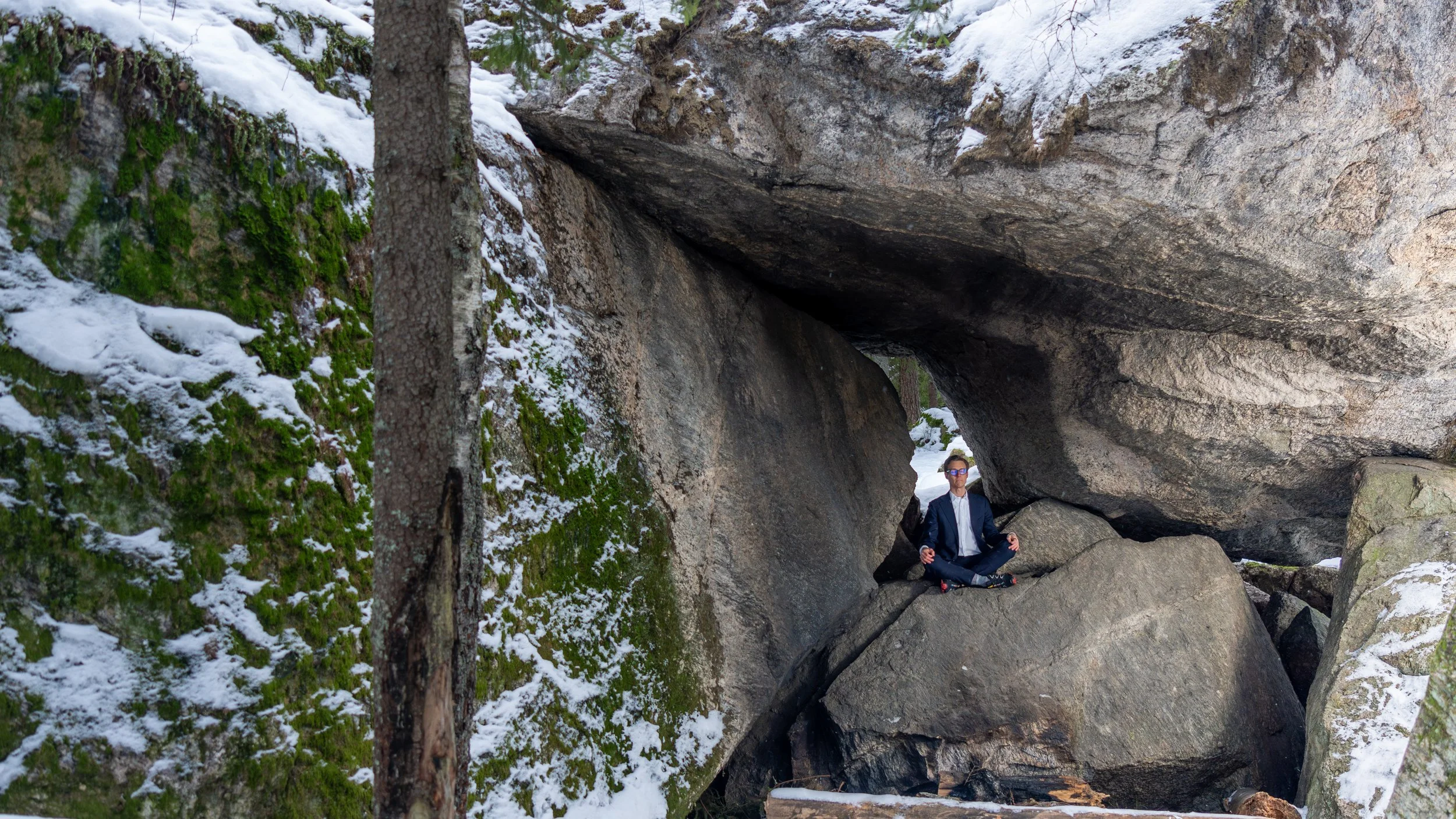 A person in a suit sitting cross-legged on a large rock under a natural rock formation in a snowy forest, with evergreen trees and snow-covered ground around.