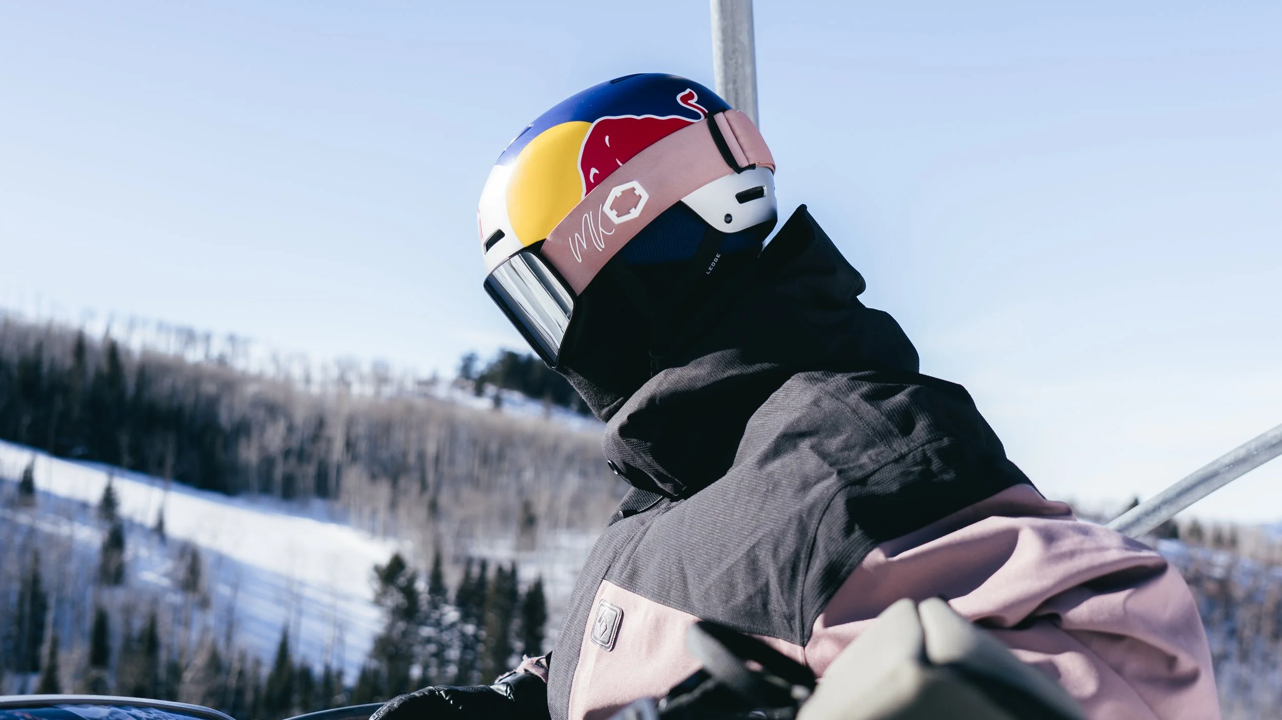 A person wearing a motorcycle helmet and winter clothing, riding a snowmobile in a snowy landscape with trees and hills in the background.