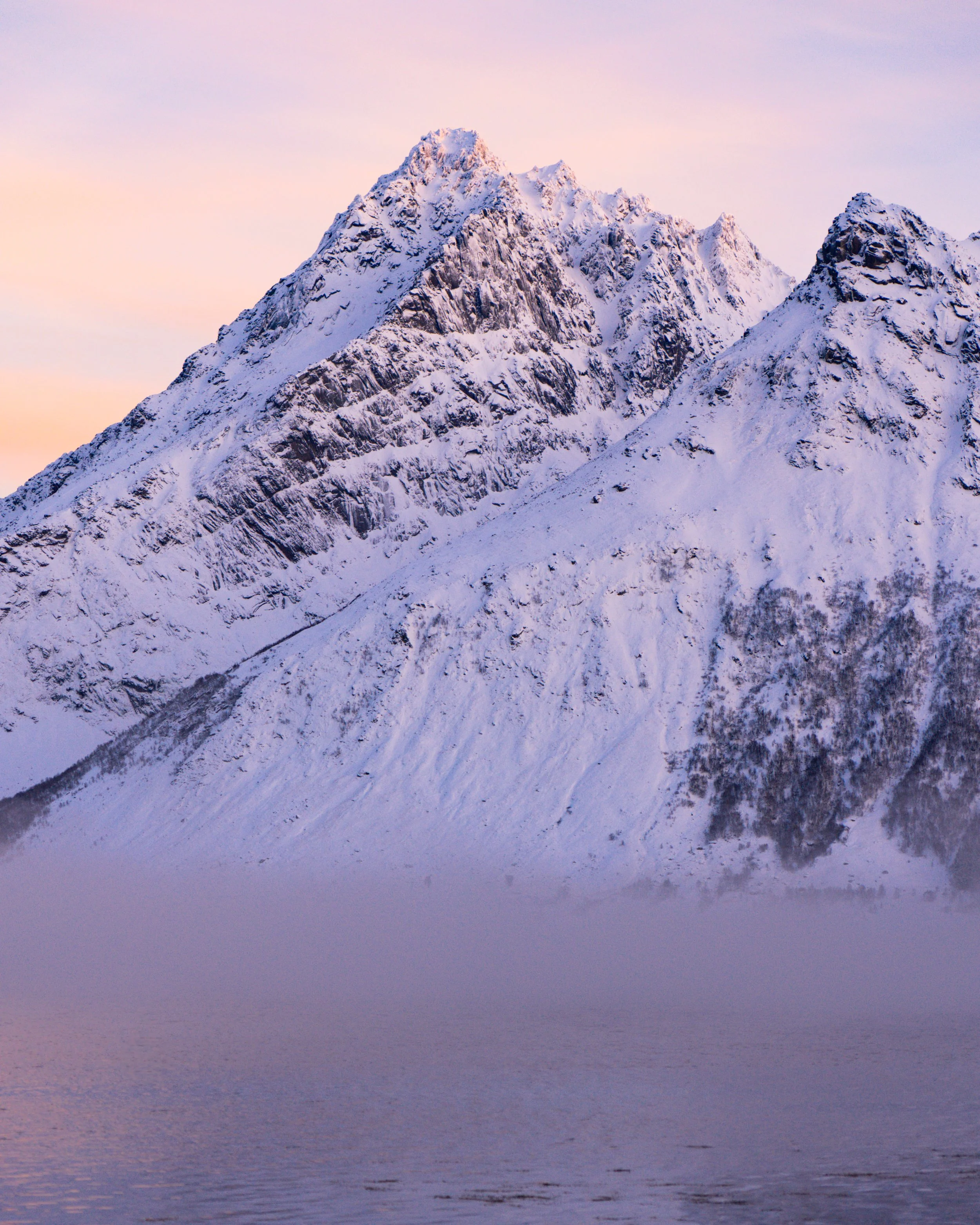 Snow-covered rugged mountain peaks at sunset or sunrise with pink and purple sky reflection.