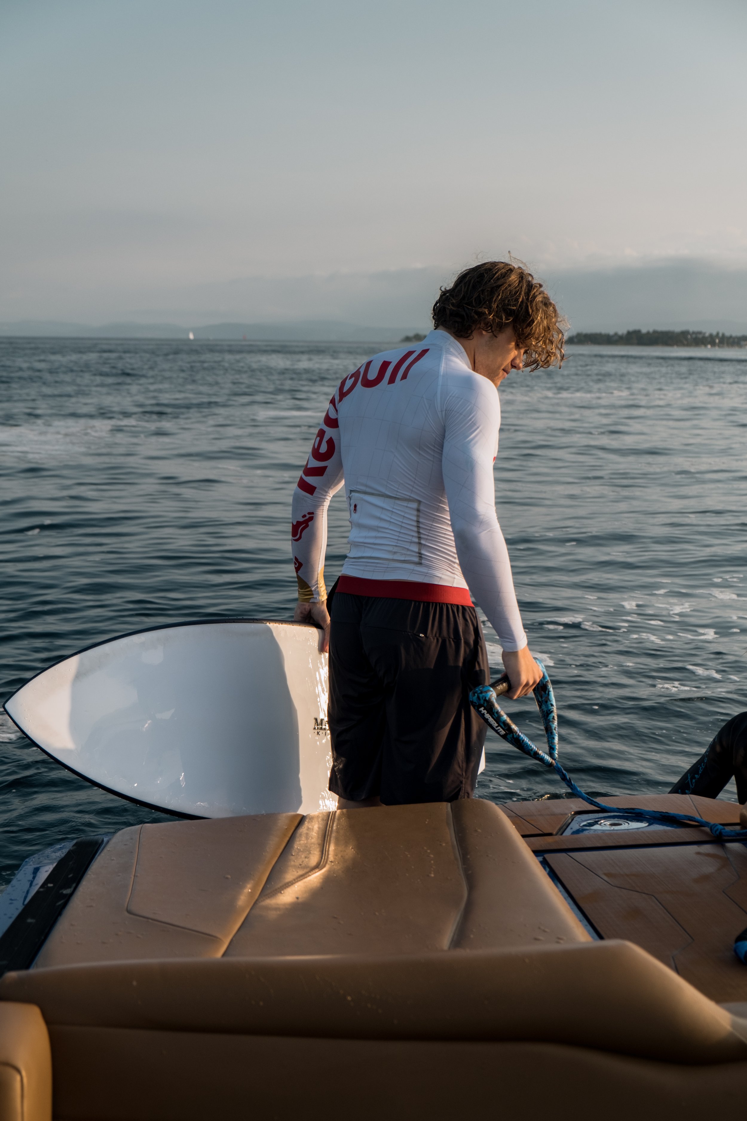 Person with curly hair in a white long-sleeve shirt and black shorts holding onto the side of a boat, preparing to surf or paddleboard near the water with a distant shoreline and sailboats in the background.