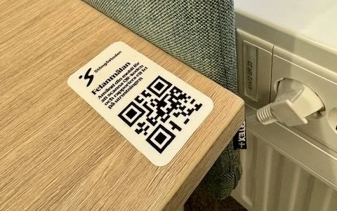 En blå telefonautomat på en väg, med en QR-kod och en felanmälan etikett på sidan.