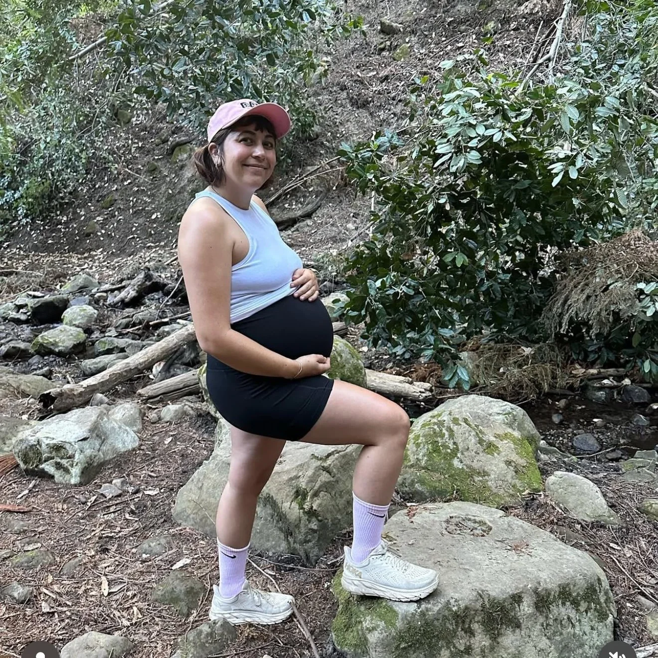 A pregnant woman with short dark hair, wearing a light pink cap, white tank top, black shorts, white socks, and white sneakers, posing on a large rock in a wooded outdoor area.