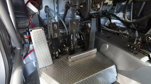 Close-up of a vehicle's interior showing racing pedals, a footrest, and a metal platform with a diamond plate pattern.