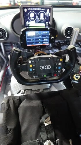 The interior of a vehicle equipped with multiple electronic devices, including a steering wheel with an Audi logo, a dashboard mounted display, and other digital screens showing various controls and information.