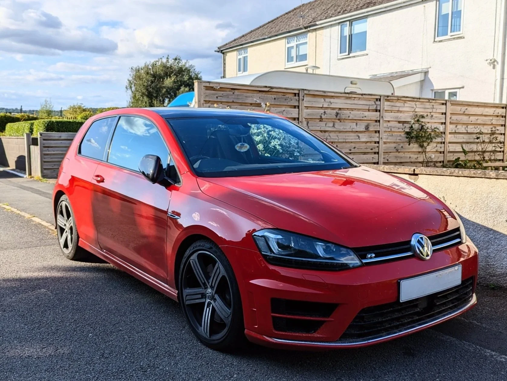 A red Volkswagen Golf R is parked on a residential street with a wooden fence and a house in the background.