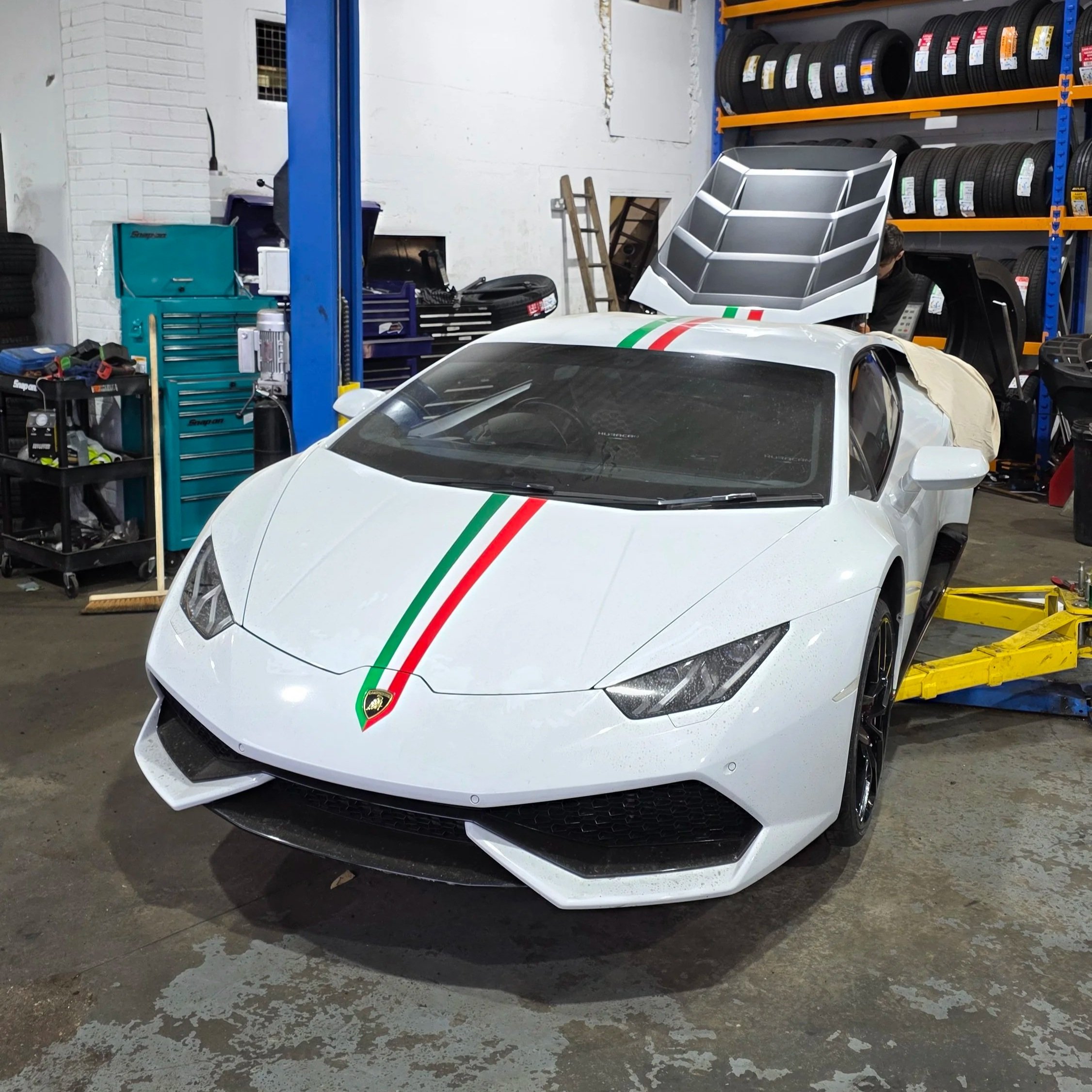White Lamborghini sports car with a green, white, and red racing stripe down the center, parked inside a garage with tires and tools.