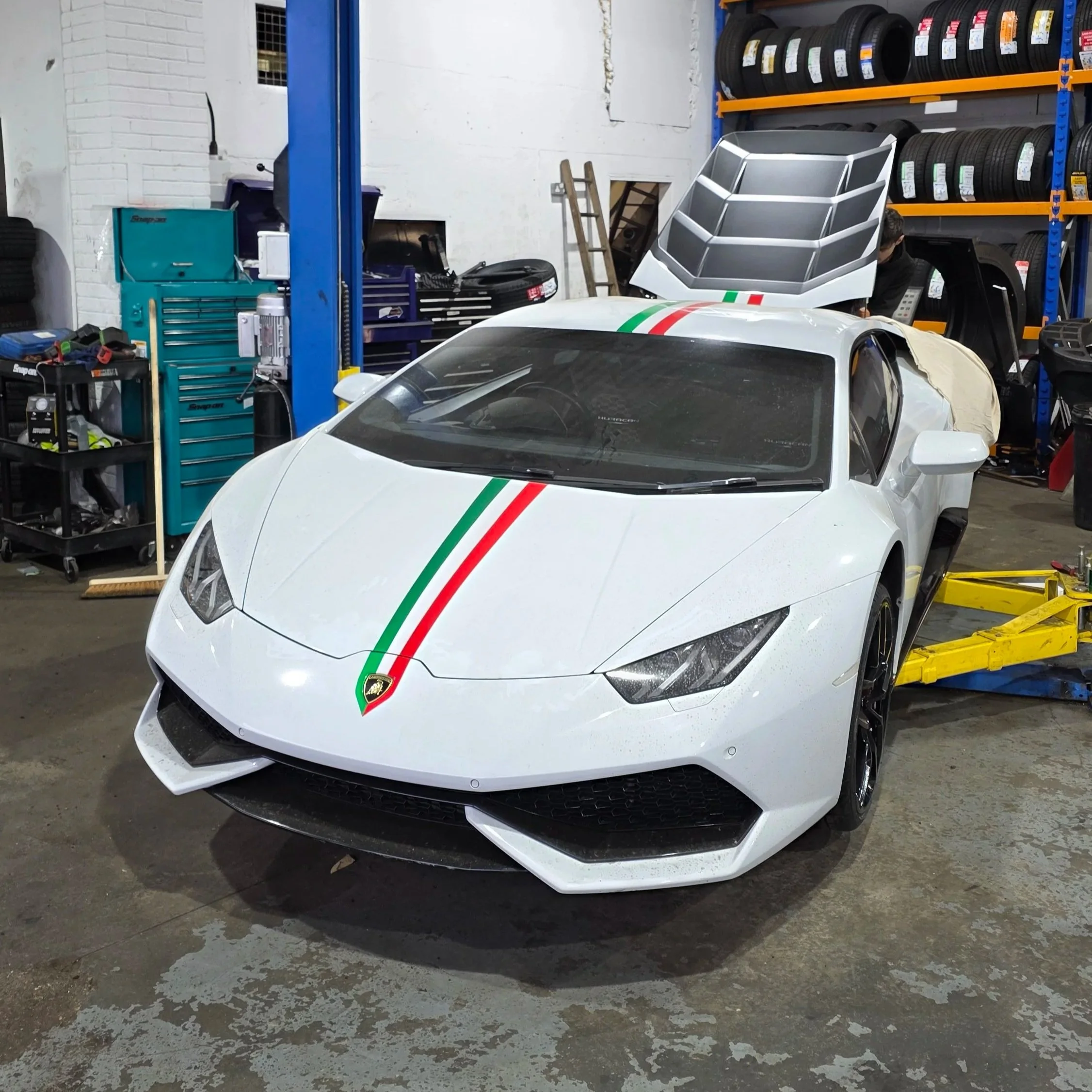 White Lamborghini sports car with Italian flag racing stripe parked inside a garage, surrounded by tools, shelves of tires, and equipment.