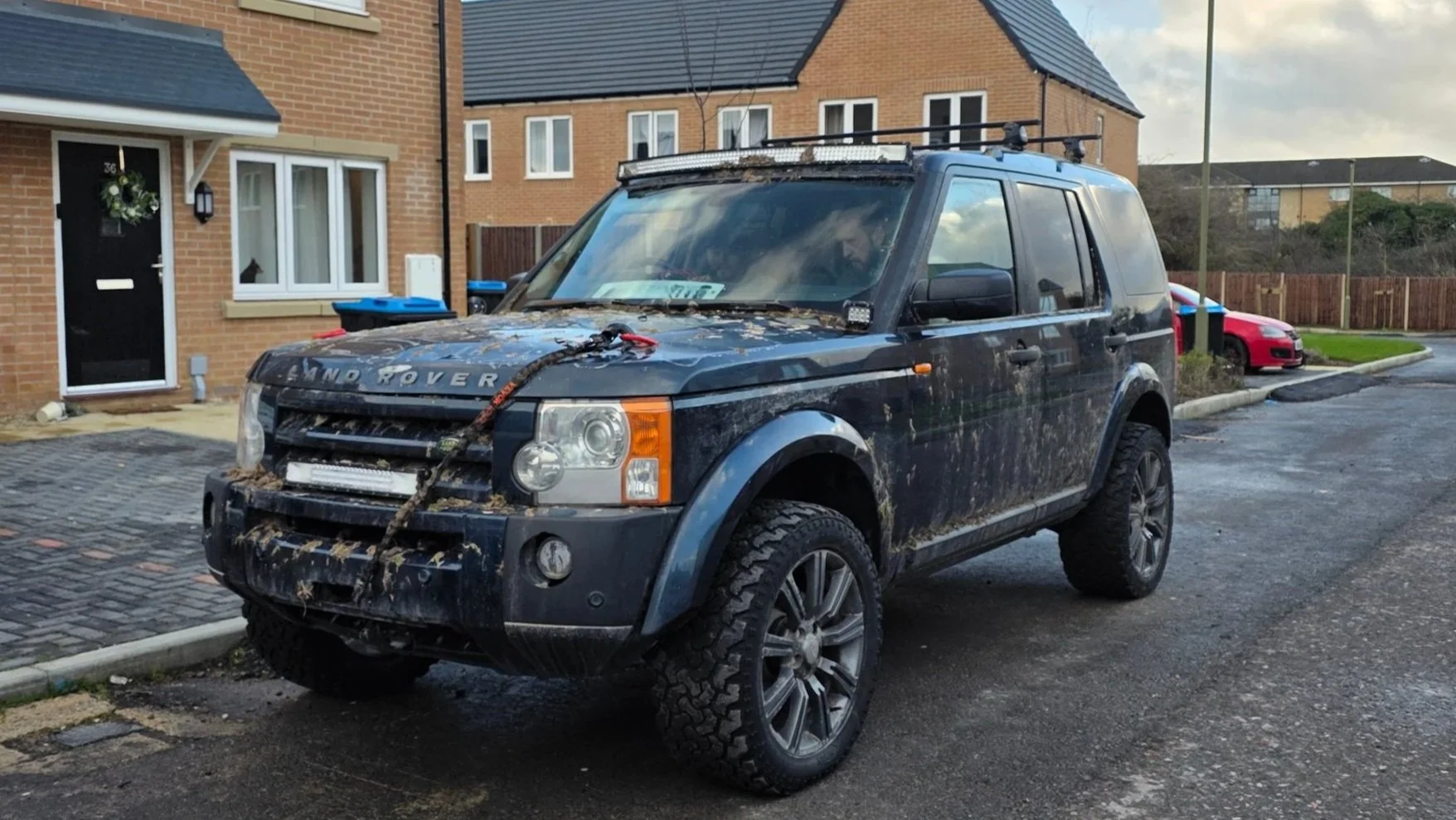 A black Land Rover parked on a residential street with mud splattered on its body and windscreen, a rope tied across the front, and mud on the tires.