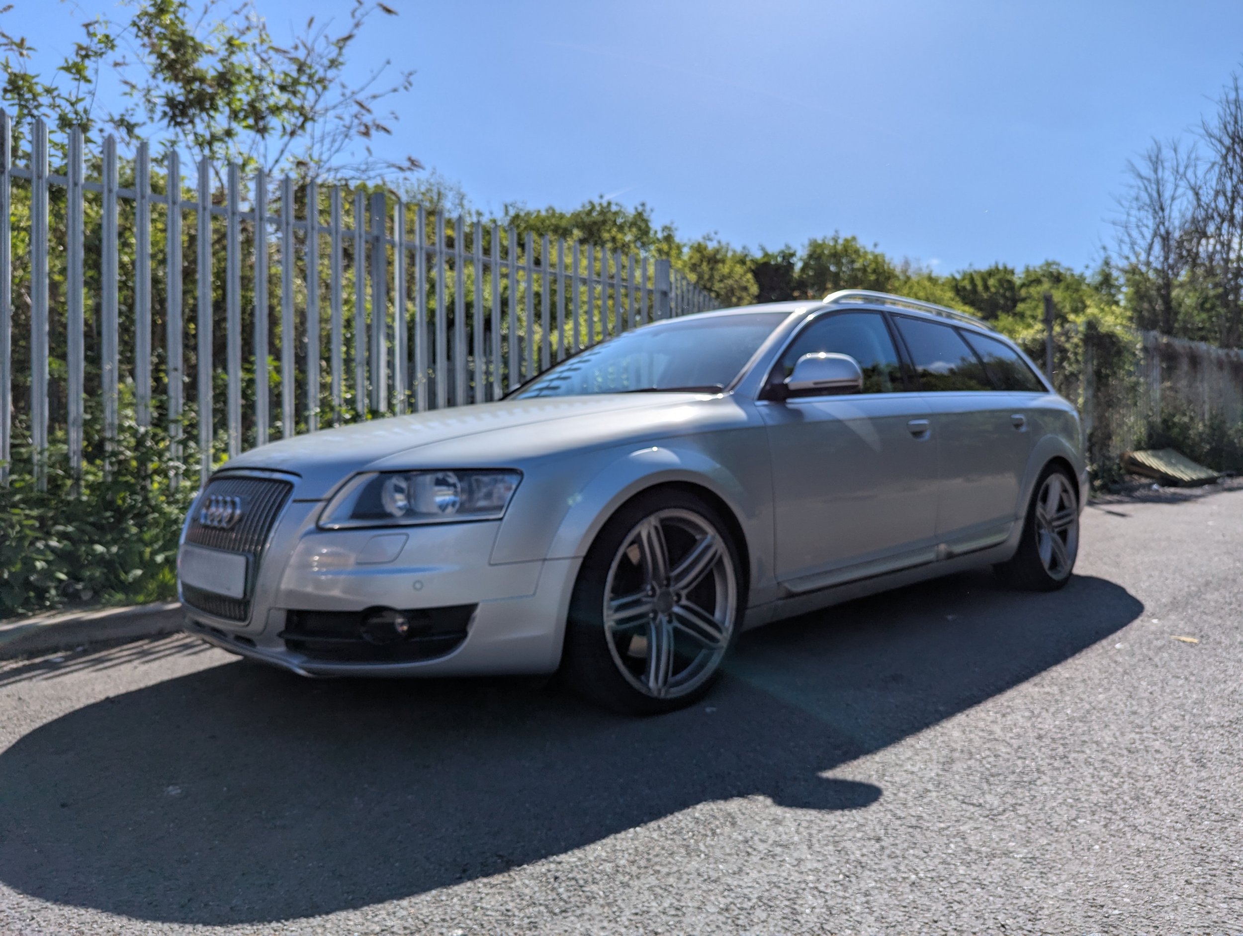Silver Audi A6 Allroad parked on a street next to a metal fence and greenery.