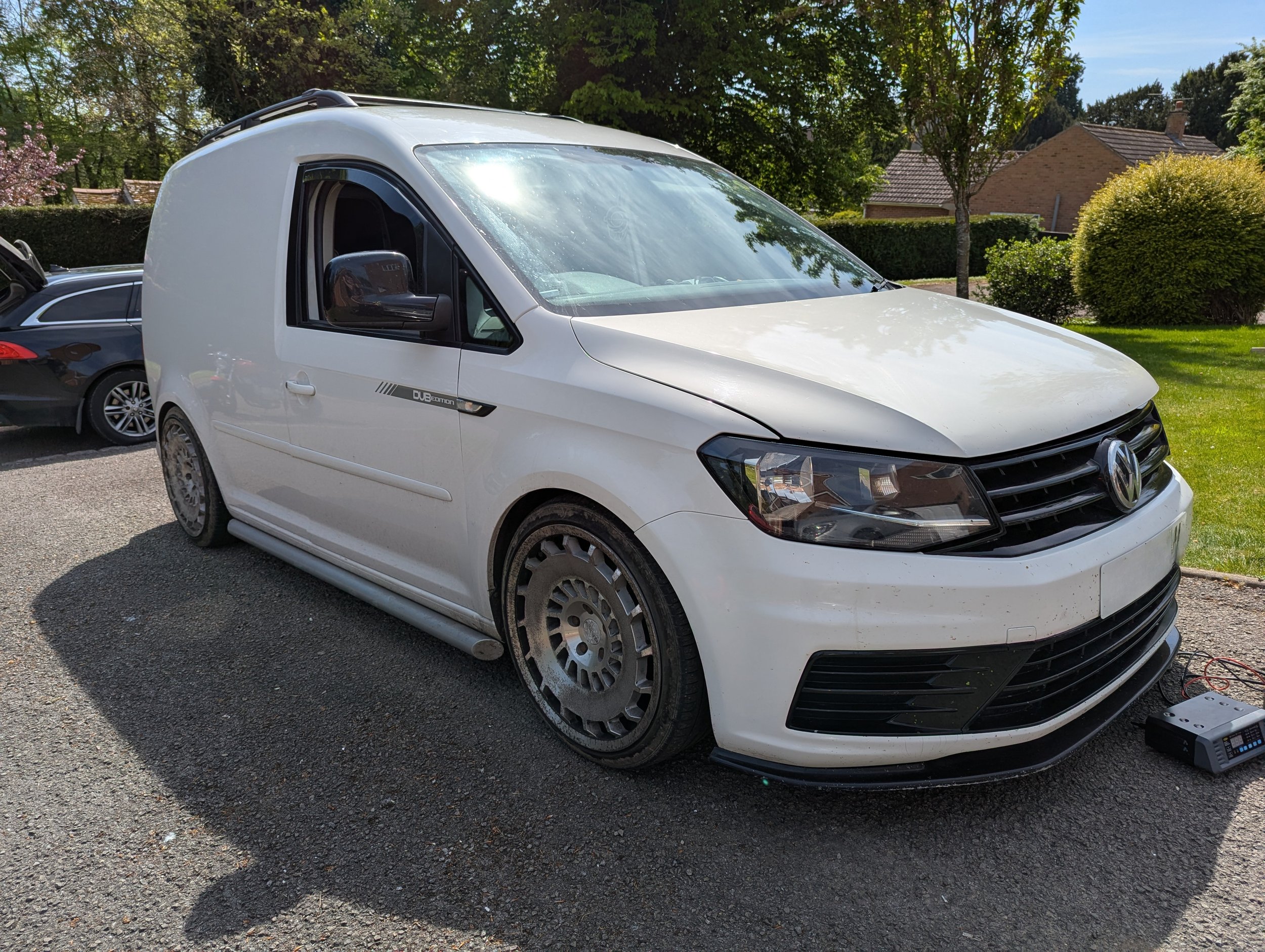 White Volkswagen Caddy van parked on a driveway, surrounded by trees and residential houses in the background.