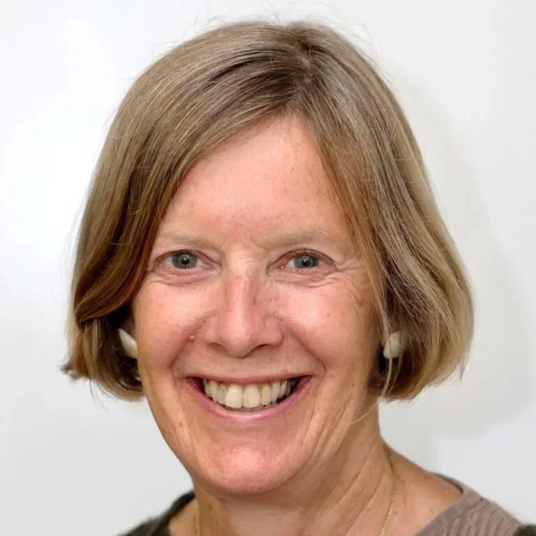 Close-up of a smiling woman with blonde hair, blue eyes, and wearing a beige top and white earrings.