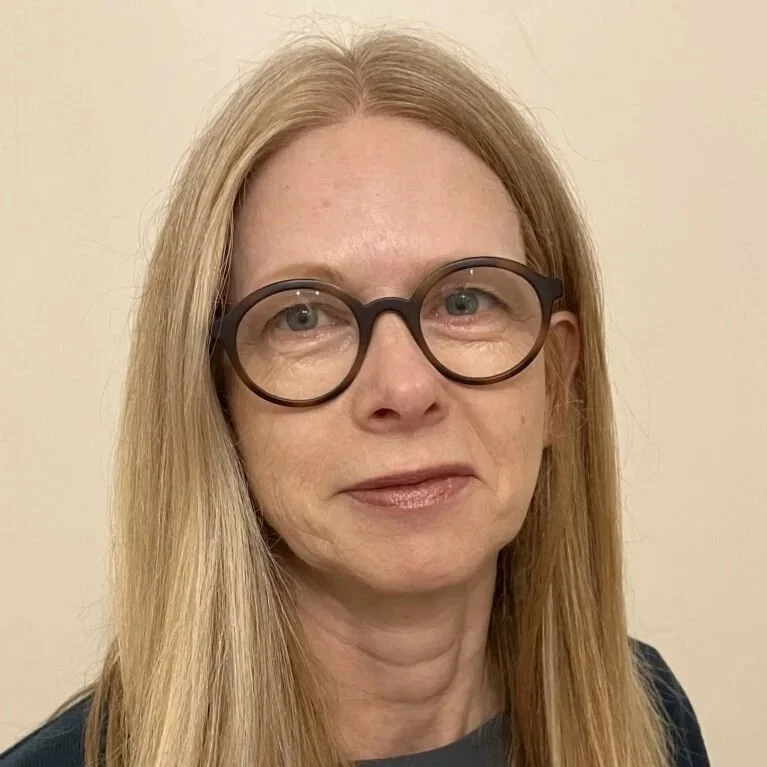 A woman with long blonde hair, wearing large round black glasses, looking at the camera, standing against a plain light-coloured wall.