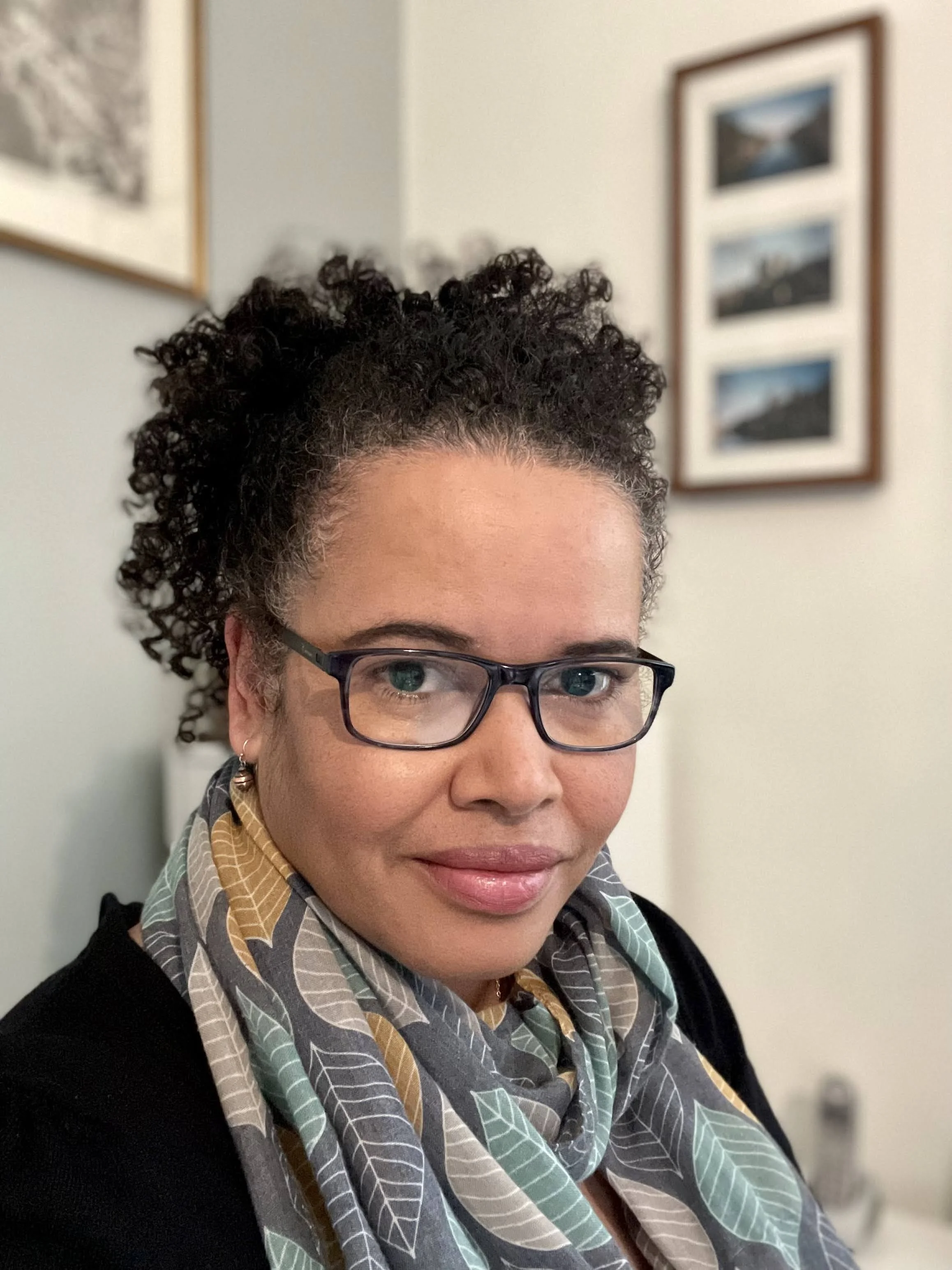 Close-up of a woman with short curly dark hair, glasses, and earrings, smiling slightly. She is wearing a patterned scarf and a dark top, with framed pictures hanging on a wall in the background.