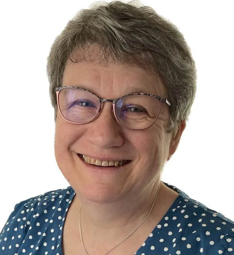 A woman with short grey hair and glasses, smiling in front of a plain white background.