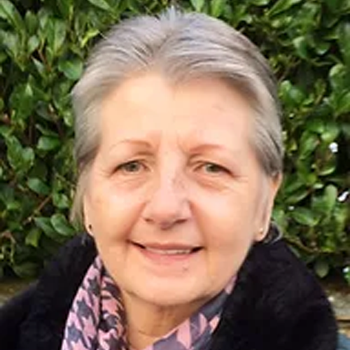 A photo of a woman with short grey hair, smiling, standing outdoors in front of green foliage.