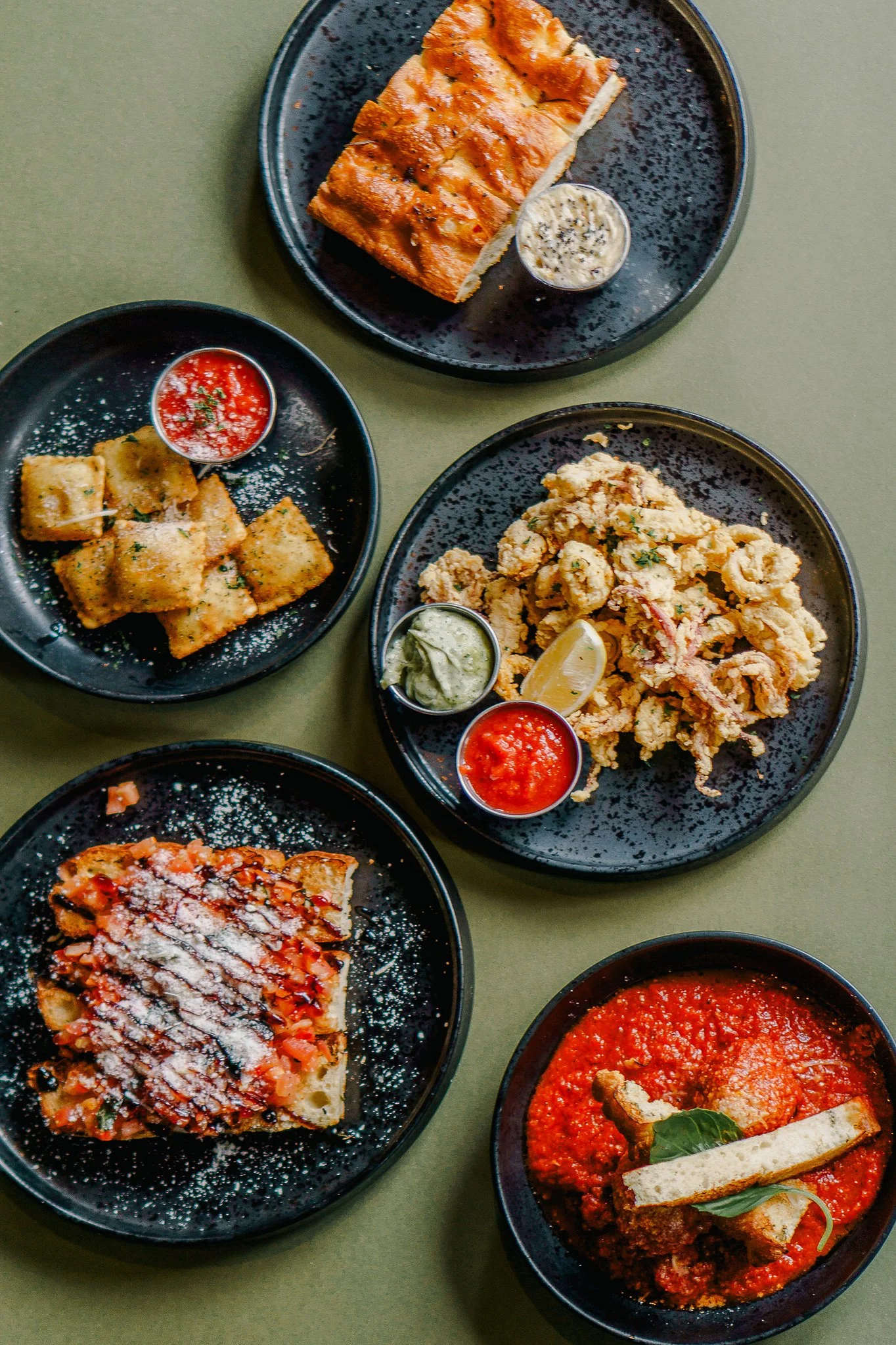 Assorted Italian dishes on black plates, including lasagna, fried calamari with dipping sauces, garlic bread, and a tomato-based stew.