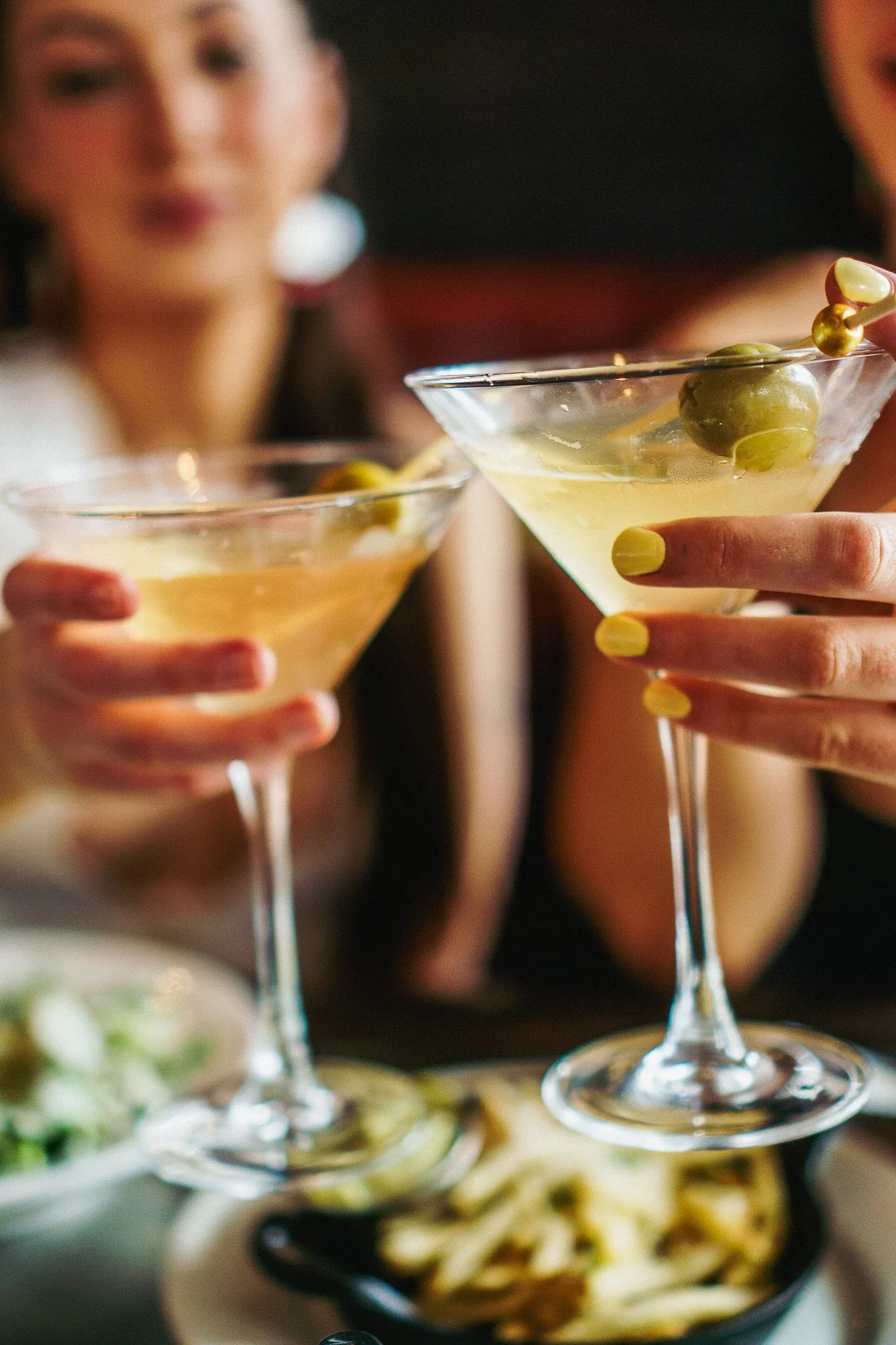 Two women holding martini glasses with green olives, with food dishes in the background.