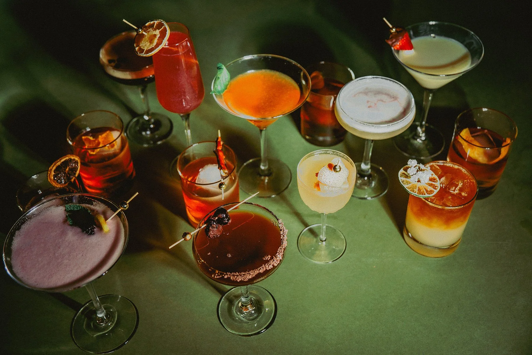 An assortment of colorful cocktails on a green surface, with various garnishes such as fruit slices, cherries, and herbs.