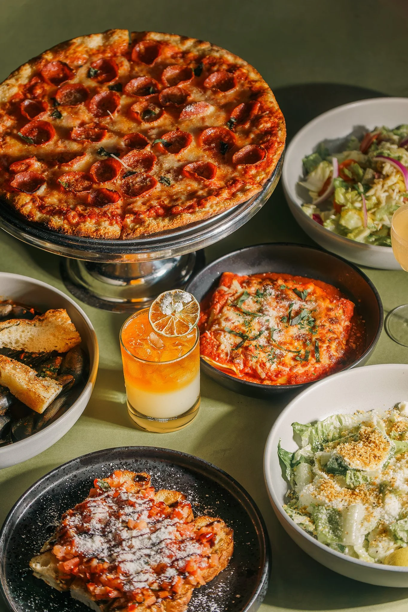 Italian food and cocktails at The Rossi Kitchen + Bar in Columbus, Ohio, featuring pepperoni pizza, baked pasta, Caesar salad, bruschetta, garlic bread, and a handcrafted cocktail.