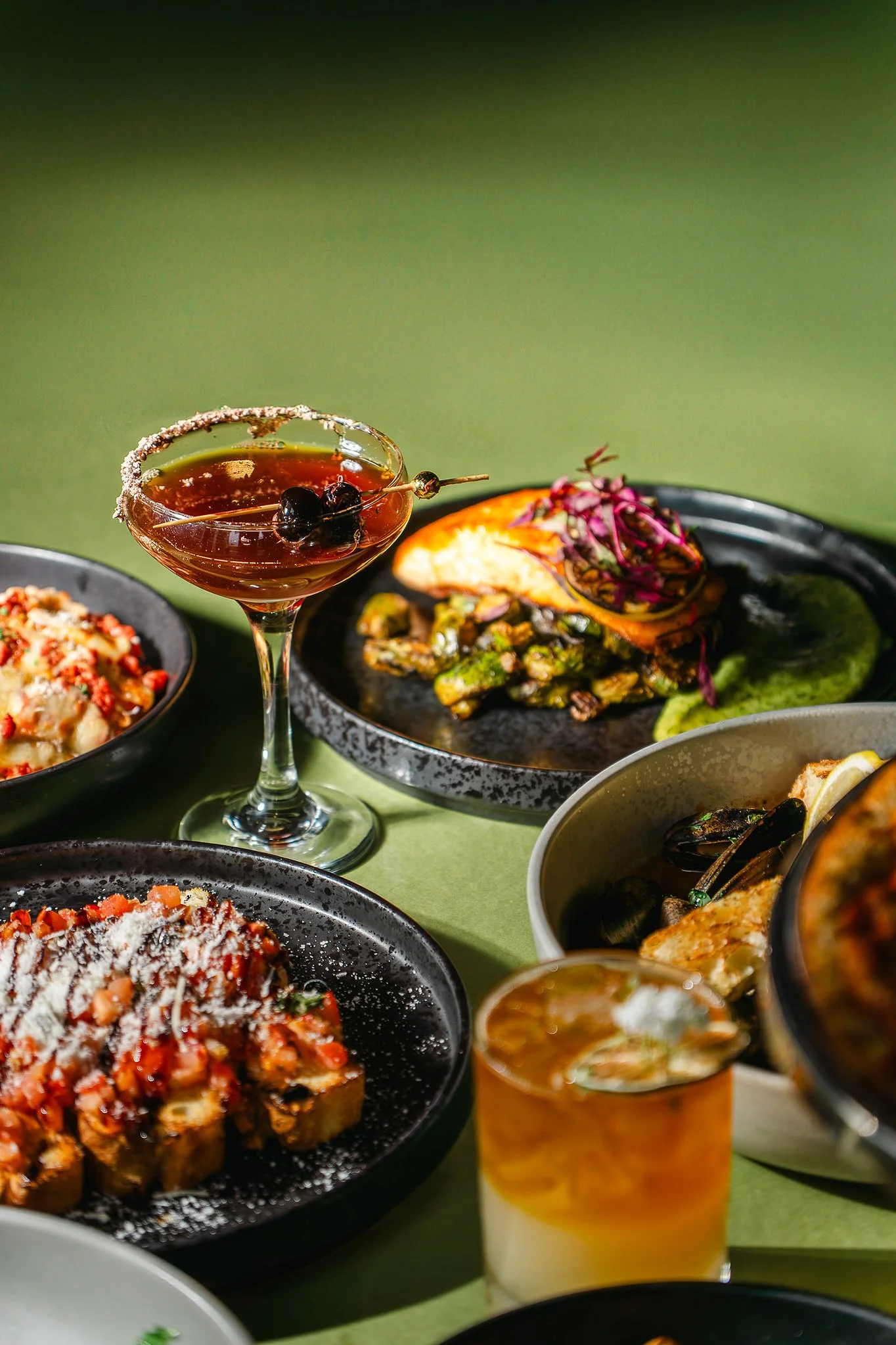 An Italian-inspired cocktail garnished with olives sits among plated dishes at The Rossi Kitchen + Bar, showcasing artfully prepared pasta, seafood, and seasonal entrées on a warmly styled table.