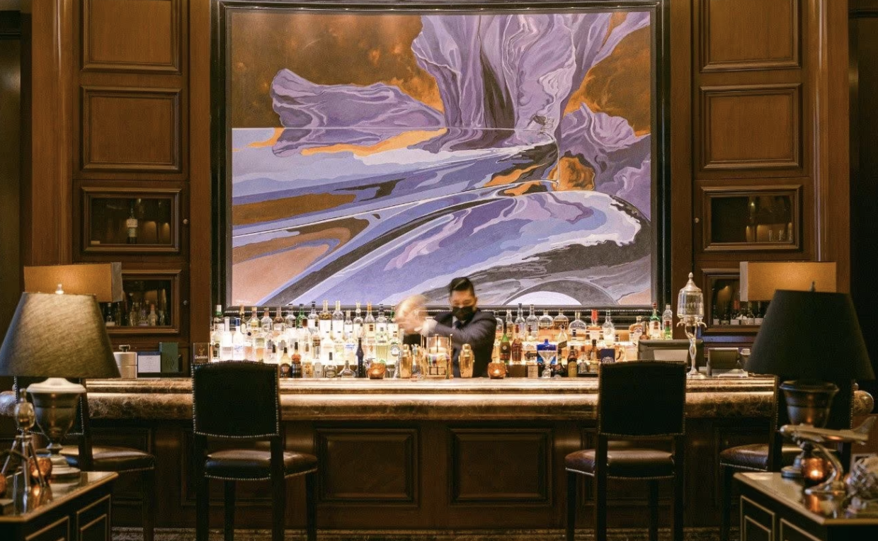 Bartender shaking cocktail behind a well-stocked bar with a large abstract painting and wooden decor in the background.