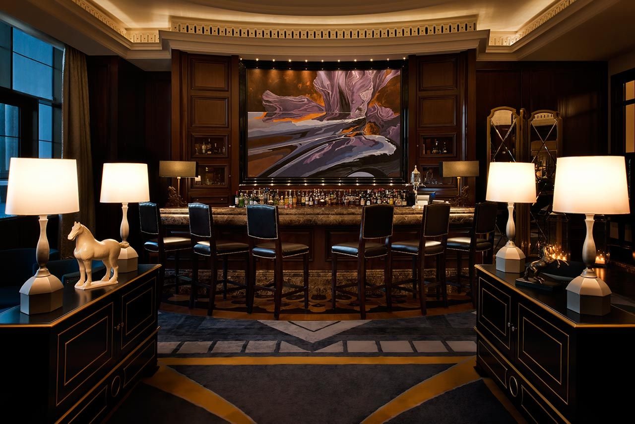 Luxury bar interior with dark wood paneling and elegant decor. Features a marble bar top lined with bottles and high-back chairs. Ambient lighting provided by table lamps with horse statues, large abstract painting as backdrop, plush carpet and curta
