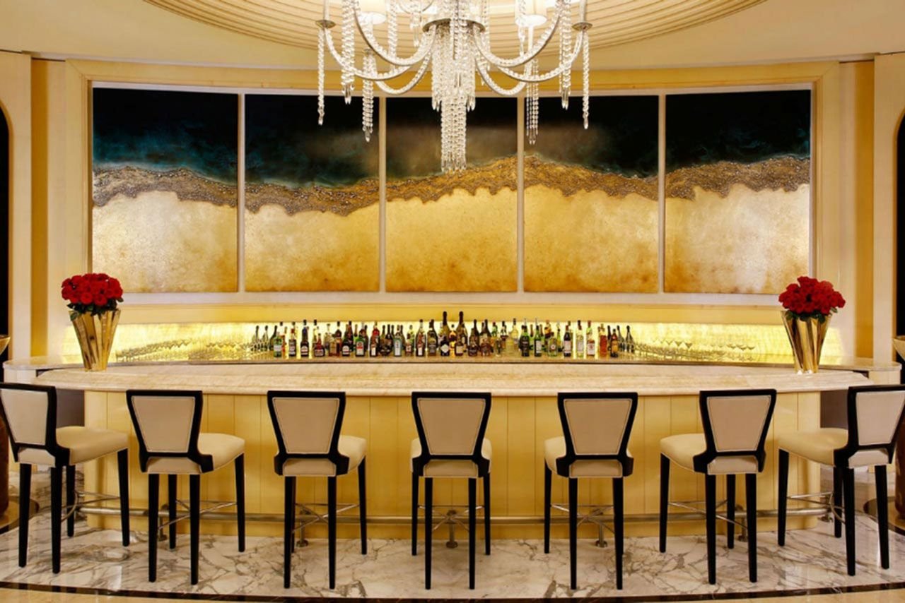 Luxurious bar interior with high chairs, marble floor, chandelier, bar counter with bottles, and decorative wall art.