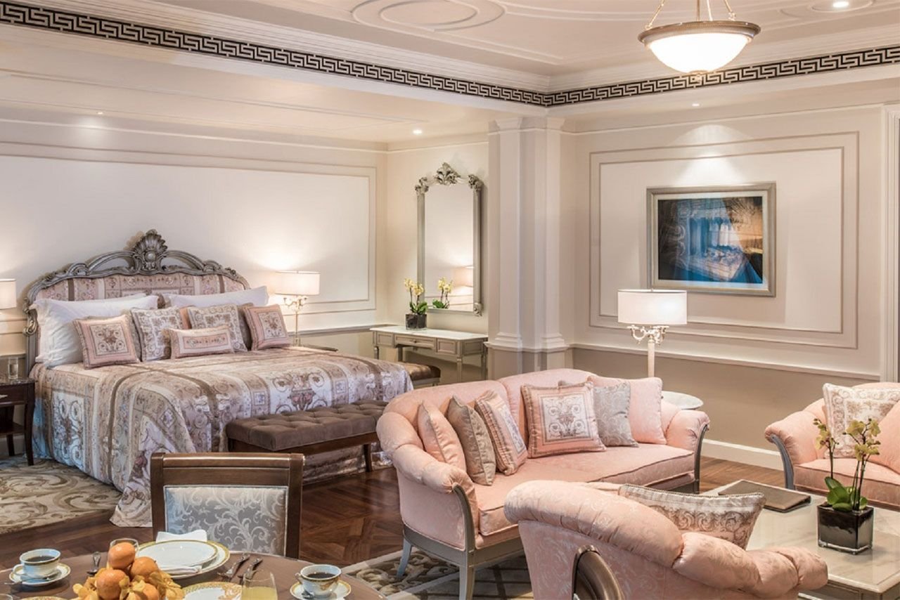 Luxurious hotel suite with king-sized bed, elegant decor, pink sofas, coffee table, wall art, and dining area.