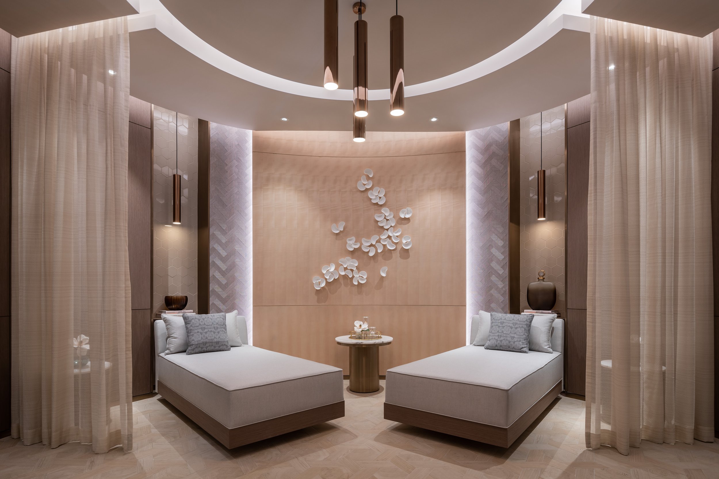 Spa relaxation room with two lounge chairs, decorative wall art, pendant lighting, beige curtains, and a small round table between the chairs.