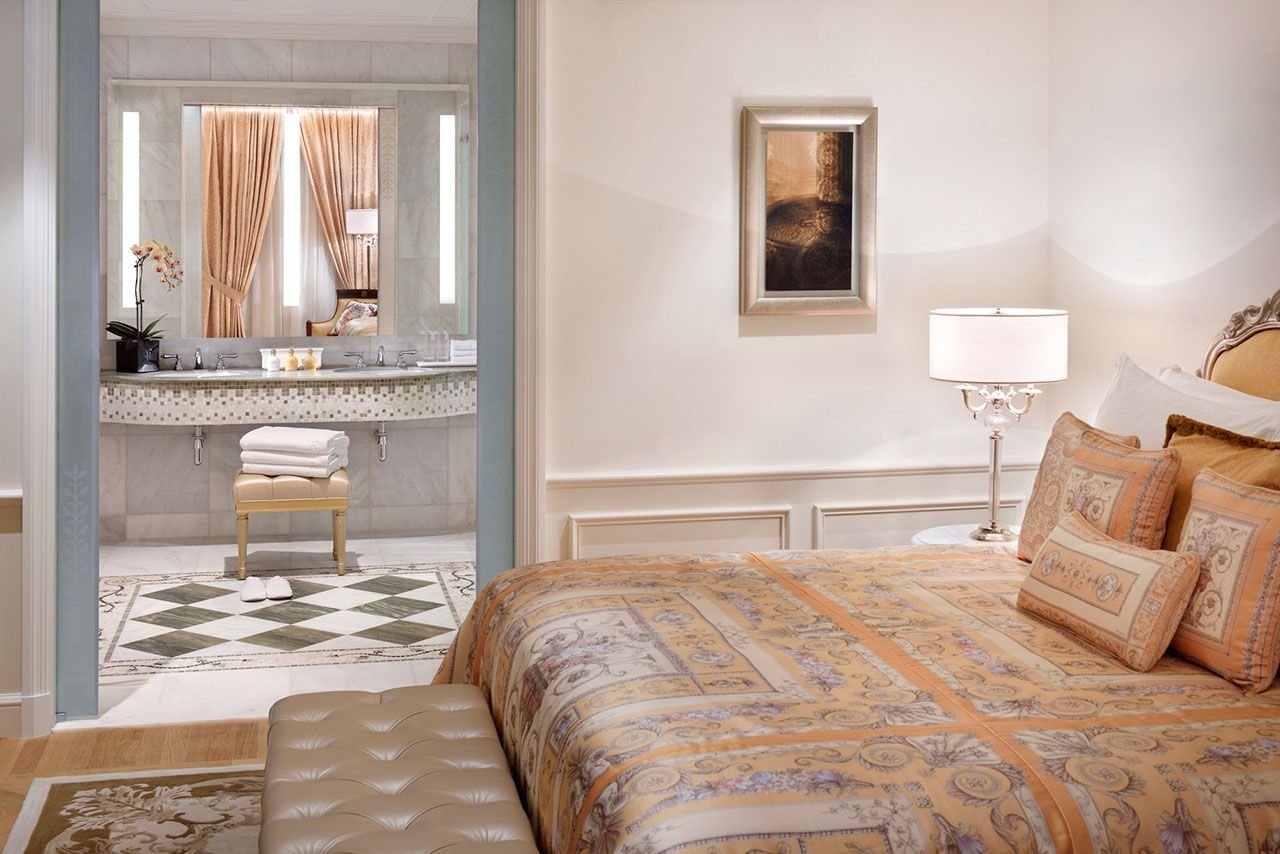 Luxurious bedroom with ornate bedding, view of marble bathroom, decorative pillows, framed artwork, and elegant lamp.