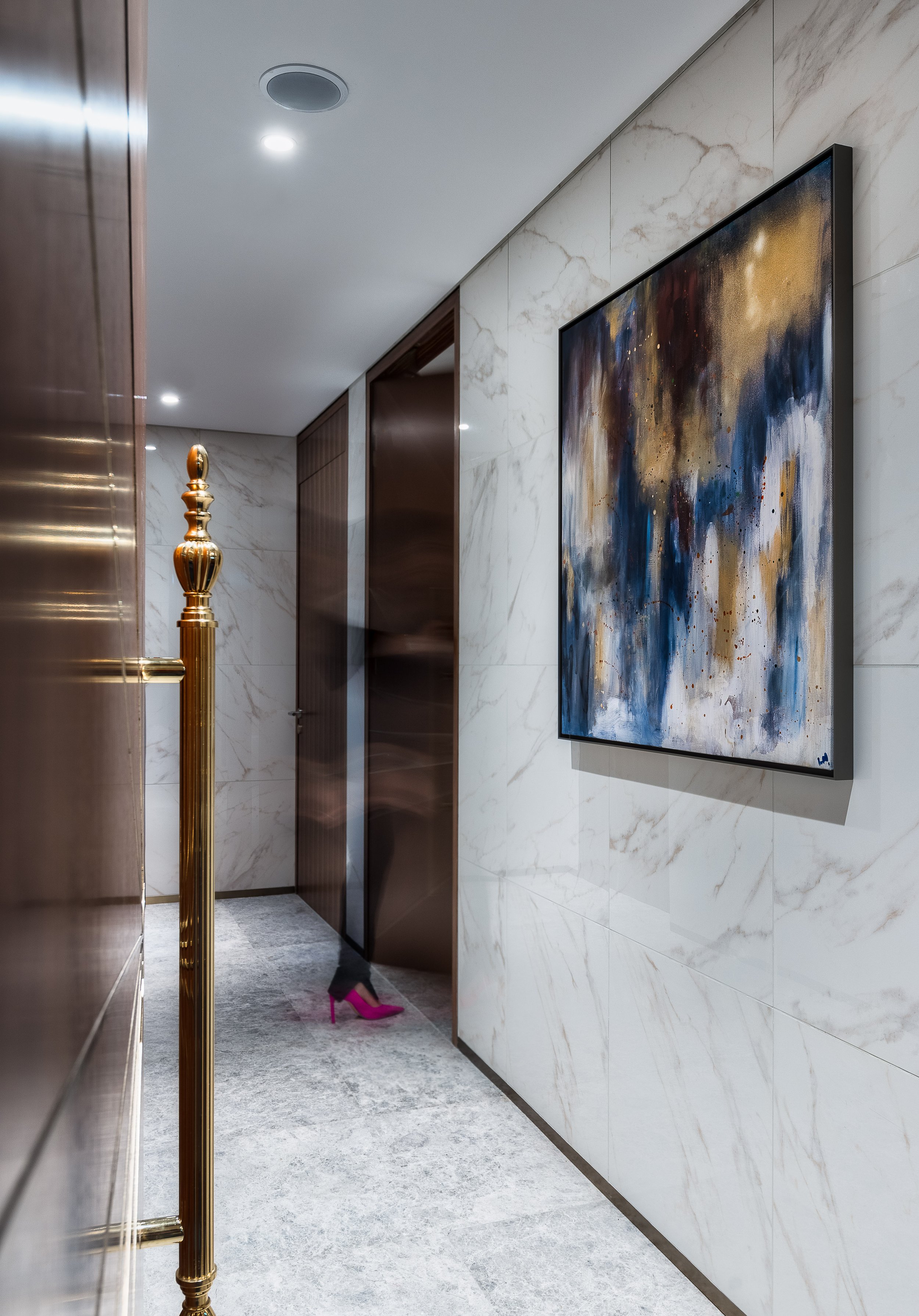 Modern hallway with abstract wall art, marble tiled walls, and a blurred figure wearing pink high heels. Brass handrail visible on the left.