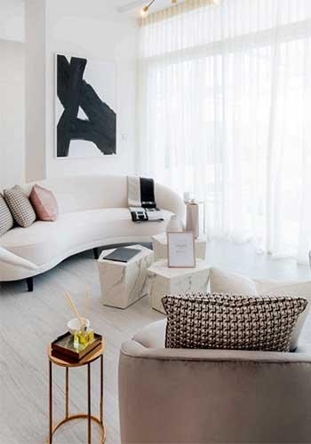 Modern living room with curved white sofa, abstract artwork, decorative pillows, small side tables, and tall windows with sheer curtains.
