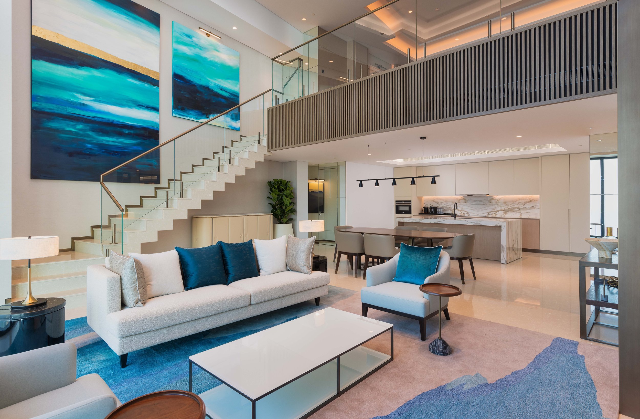 Modern living room and kitchen with staircase, abstract wall art, beige and blue furniture, glass railing, and open layout.