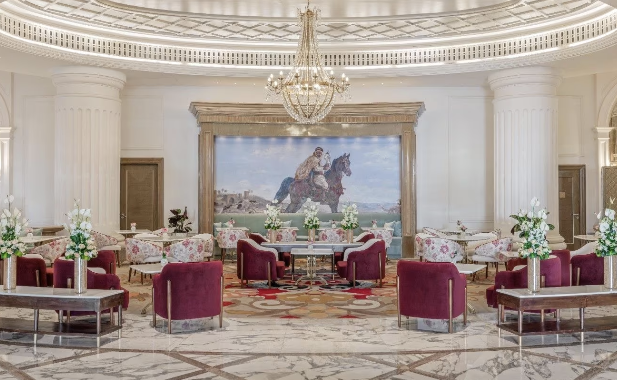 Opulent hotel lobby with chandelier, large painting of horse and rider, red and white chairs, and floral arrangements.