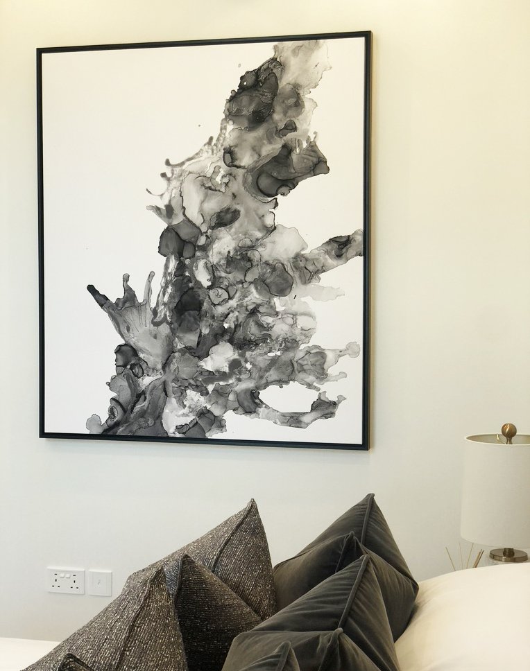 Abstract black ink painting on white canvas, framed on a wall above a bed with dark gray pillows and a bedside lamp.