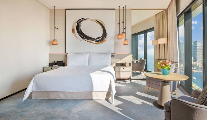 Modern hotel room with king-size bed, abstract wall art, large windows with city view, and contemporary furniture.