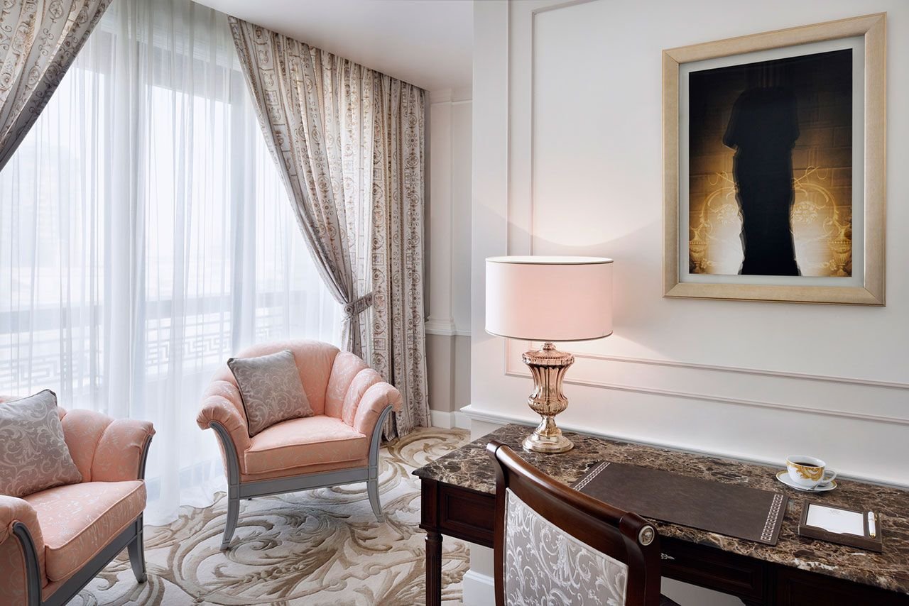 Elegant sitting area with two pink upholstered chairs, a decorative desk with marble top, table lamp, framed wall art, and ornate curtains with sheer panels.