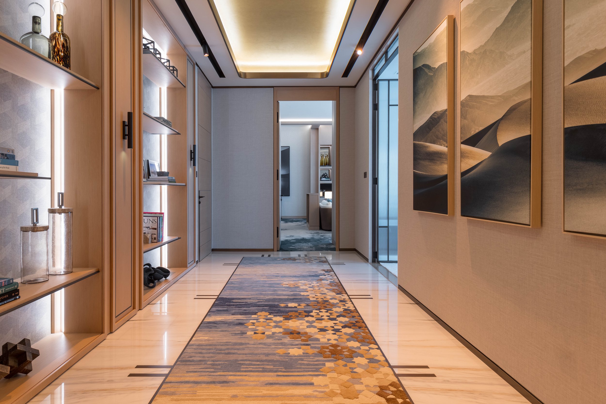 Modern hallway with built-in wooden shelves, decorative vases, books, and wall art. A long rug with geometric patterns covers the floor leading to a doorway.