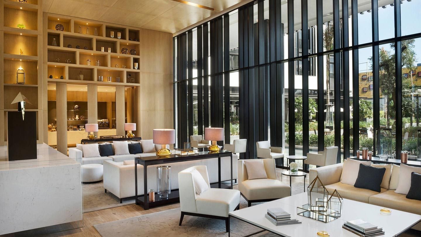 Modern hotel lobby with a spacious seating area featuring white sofas and armchairs, large black floor-to-ceiling windows, decorative shelving, and lighting fixtures on tables.
