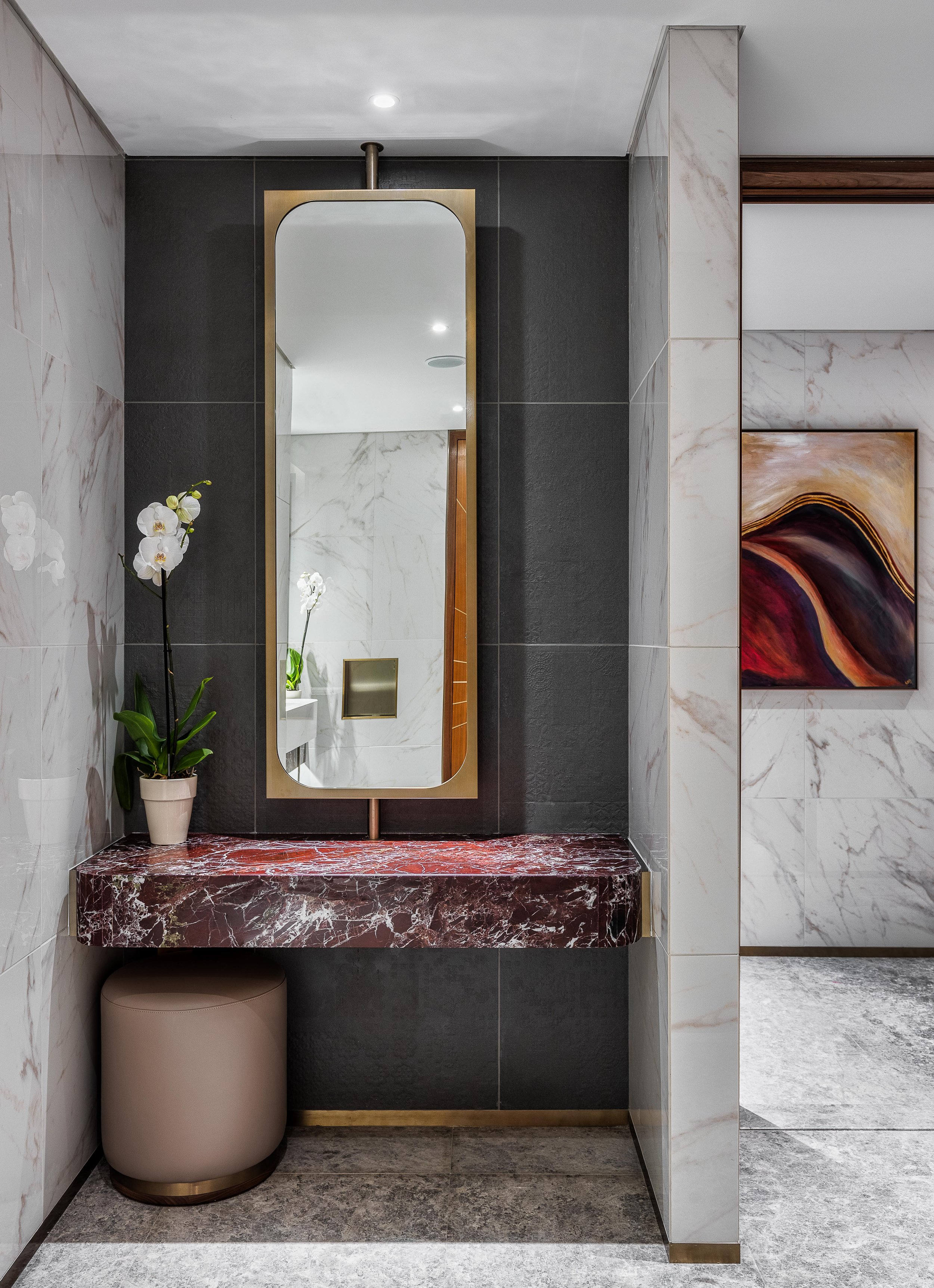 Modern bathroom vanity with rectangular mirror, red marble countertop, potted orchid, round stool, and abstract wall art.