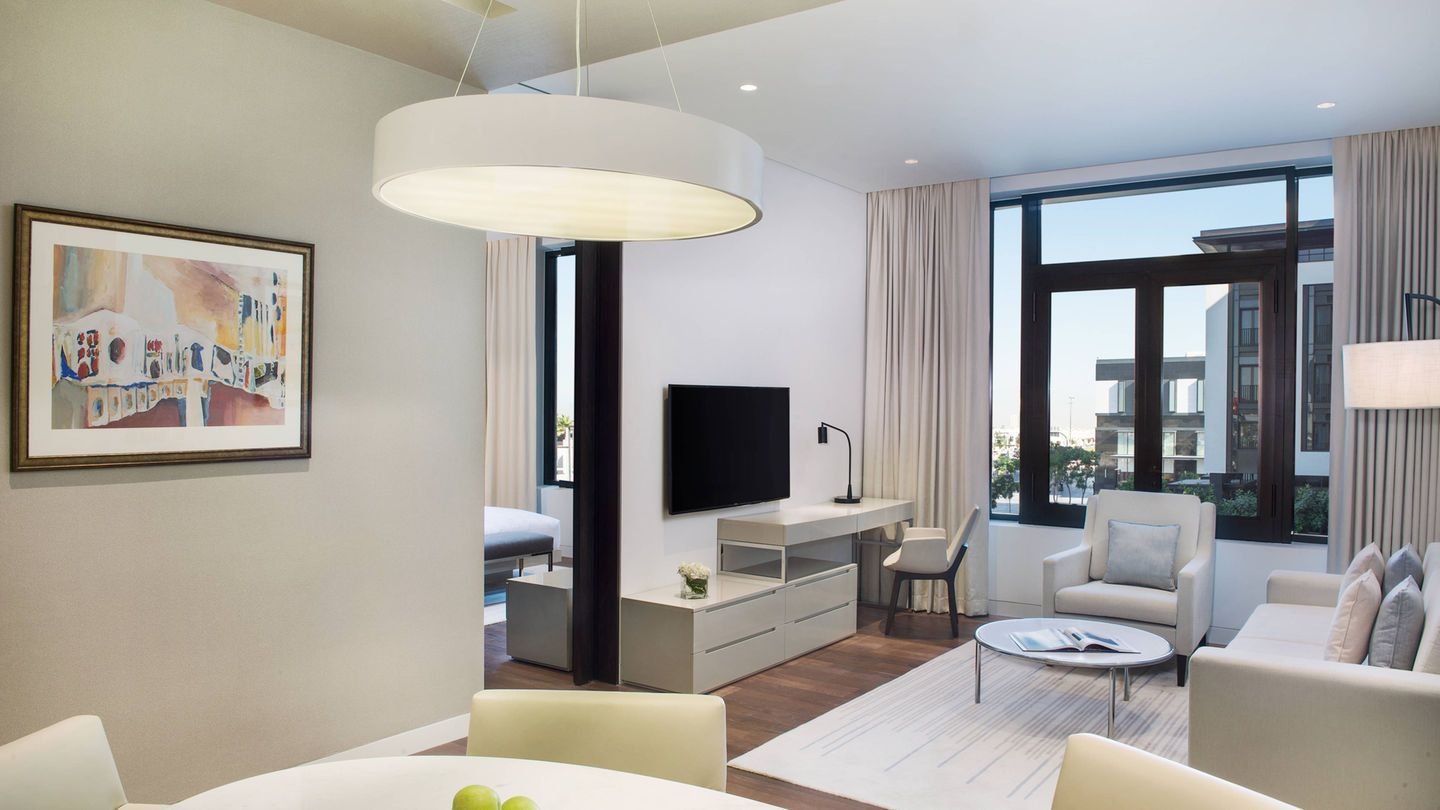 Modern living room with large windows, white walls, and wooden floor. Features a cream sofa, armchair, round coffee table, wall-mounted TV, desk with chair, abstract painting, and pendant light. Apples on a dining table in the foreground.