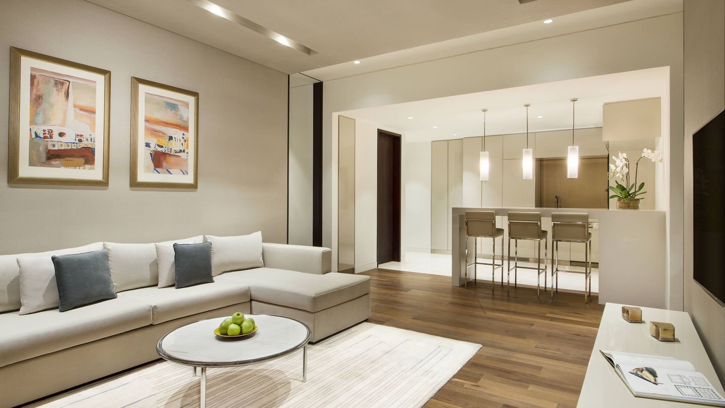 Modern living room with beige sectional sofa, abstract art, round coffee table with green apples, open kitchen with bar stools, pendant lights, and hardwood flooring.
