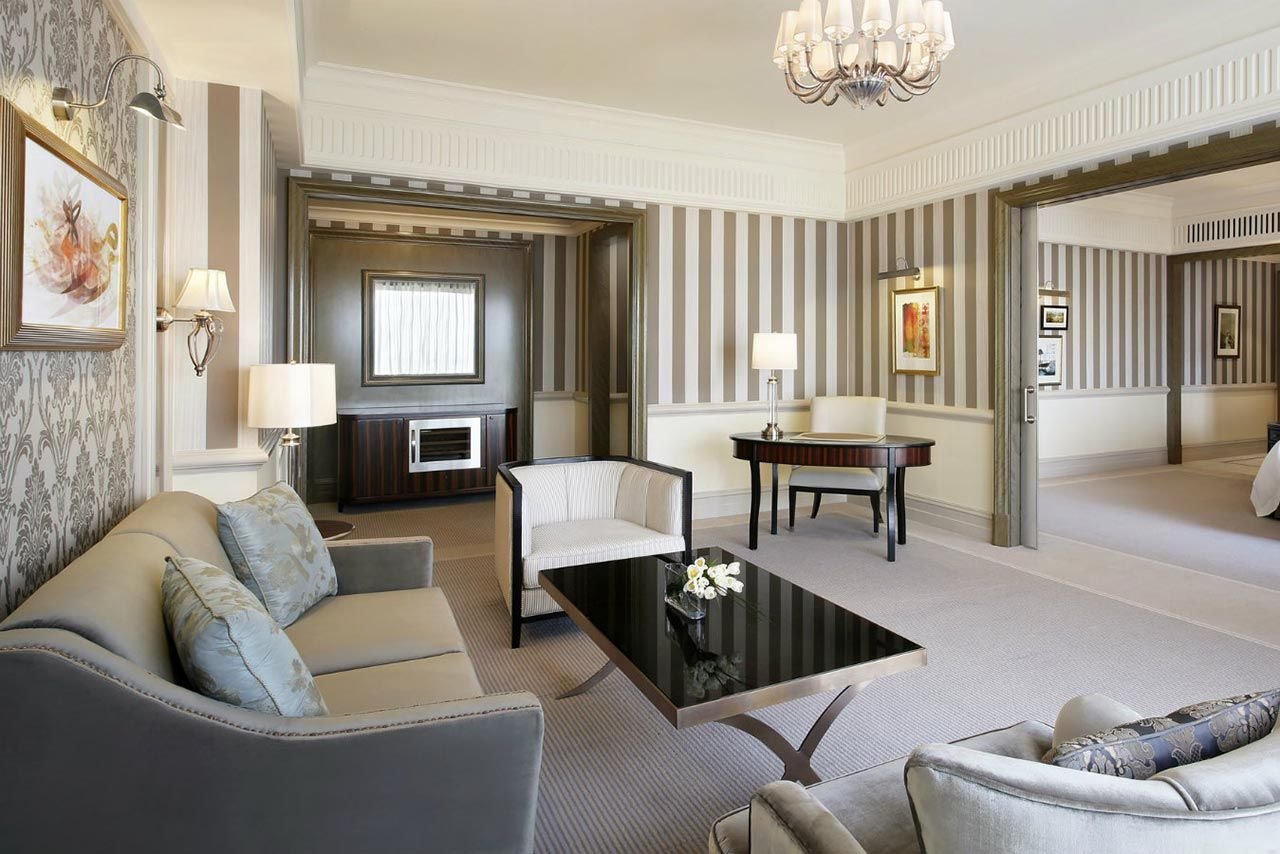 Luxurious hotel living room with elegant furniture, striped wallpaper, a modern coffee table, sofa, armchair, desk, and wall art.