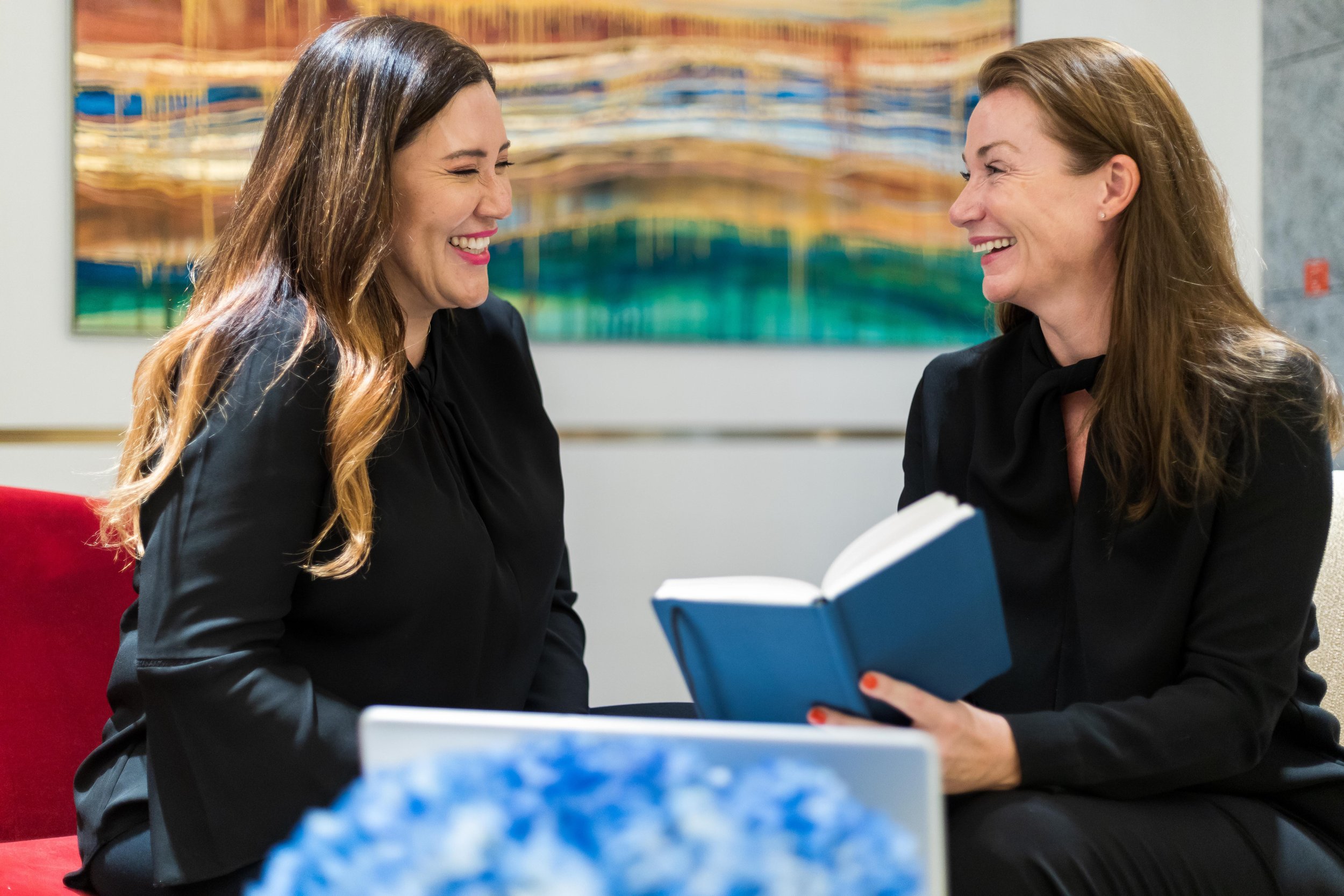 Two women sitting and laughing with a book in hand, in front of a colorful abstract painting.