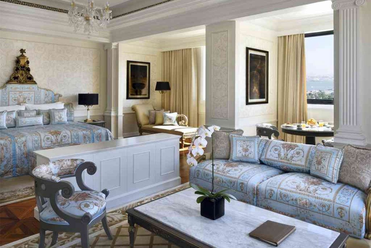 Elegant hotel suite with luxurious decor, featuring a king-sized bed, plush seating, ornate furnishings, marble coffee table with orchid plant, and a small dining area by a window overlooking a cityscape.