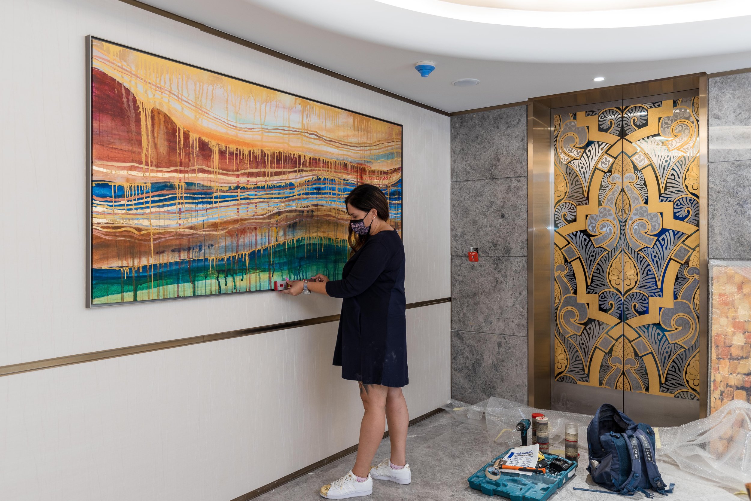 Woman adjusting modern abstract painting on wall in a room with ornate elevator doors and a toolbag on the floor.